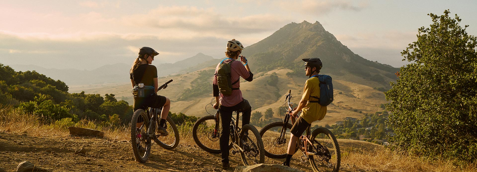 three mountain bikers take a break on a dirt trail overlooking rolling hills and a prominent mountain peak in golden evening light