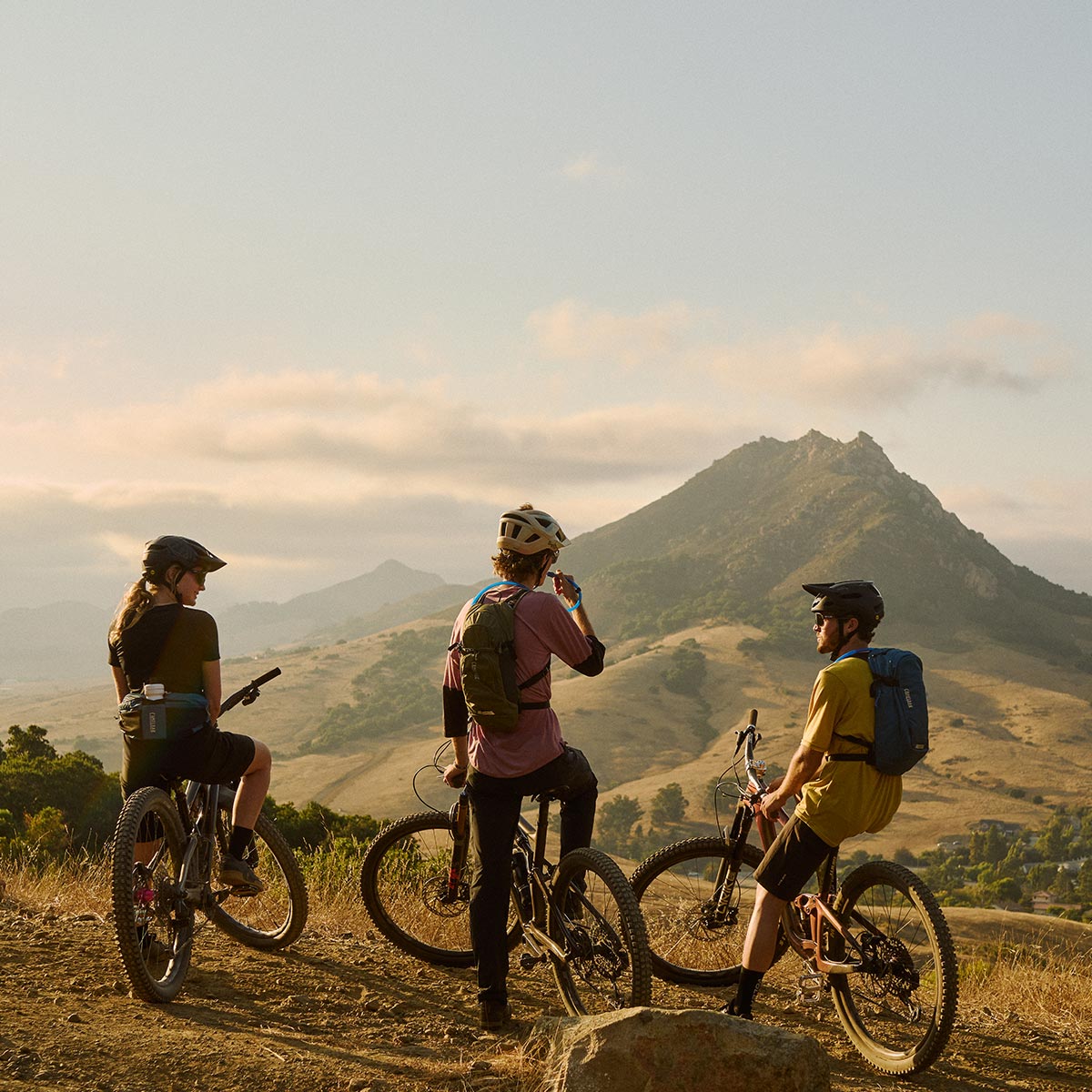three mountain bikers take a break on a dirt trail overlooking rolling hills and a prominent mountain peak in golden evening light
