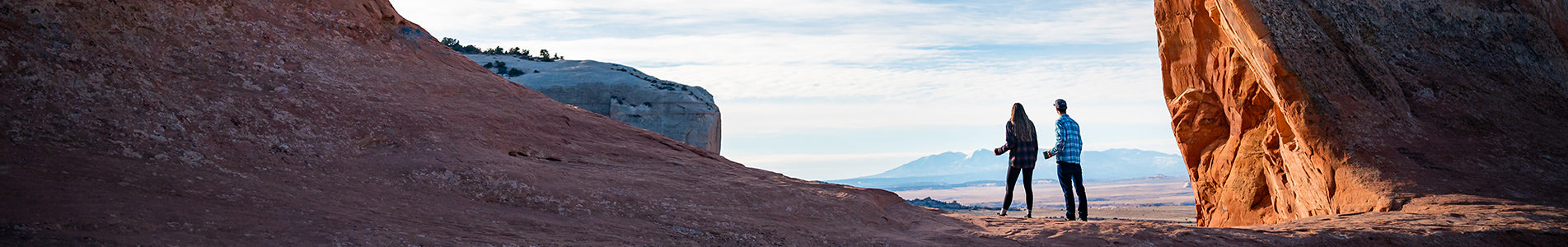 two people explore a natural stone arch or cave opening in red rock country, silhouetted against an expansive desert valley and mountain range